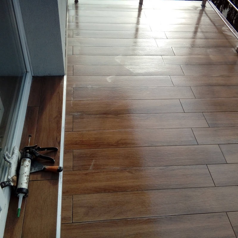 Ułożone brązowe płytki imitujące drewno na balkonie, widoczny pistolet do silikonu i szpachelka przy ścianie.