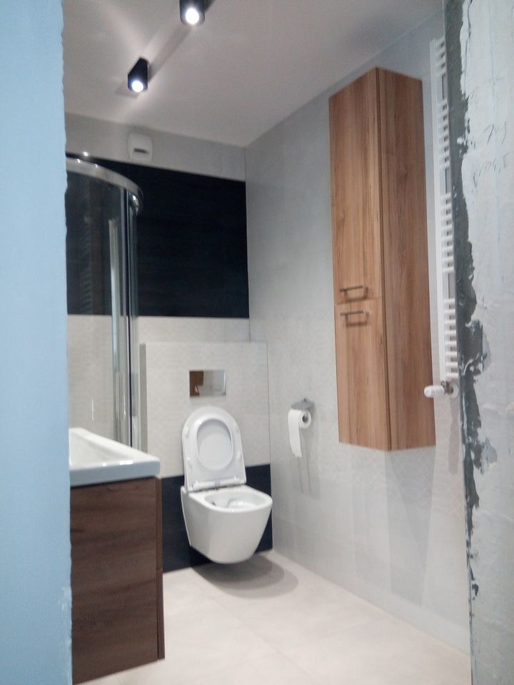 Nowoczesna łazienka w trakcie wykańczania: podwieszana toaleta z otwartą klapą, szafka pod umywalką w kolorze drewna, wysoka szafka wisząca z drewnianym fornirem i grzejnik drabinkowy na ścianie...