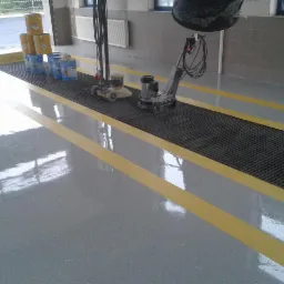 Świeżo wylana, błyszcząca posadzka żywiczna w odcieniach szarości z żółtymi pasami, z maszynami do polerowania i wiaderkami z materiałami na kratce ściekowej.