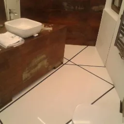 Nowoczesna łazienka z białą posadzką z czarnymi liniami, umywalką nablatową na szafce z efektem postarzanego drewna oraz grzejnikiem drabinkowym na białej ścianie.