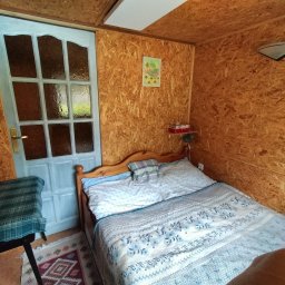 Mały Domek na Kaszubach - Drewniany pokój gościnny z łóżkiem dwuosobowym, stolikiem i jasnoniebieskimi drzwiami. Ściany wyłożone płytami OSB, dekoracje na ścianie. Przytulne wnętrze.