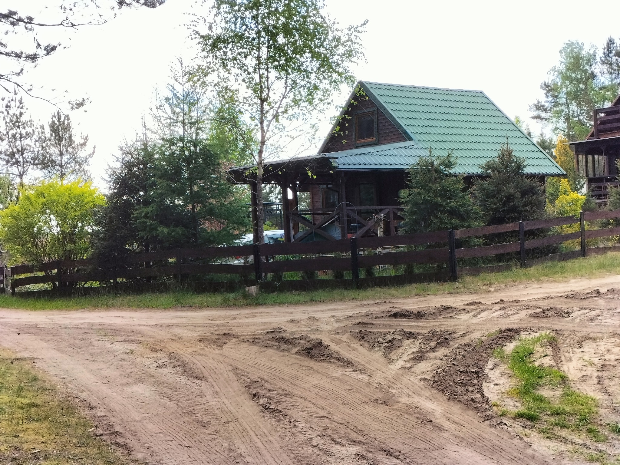 Drewniany domek letniskowy z zielonym dachem, otoczony drzewami i drewnianym płotem. Piaszczysta droga dojazdowa z wyraźnymi śladami opon.