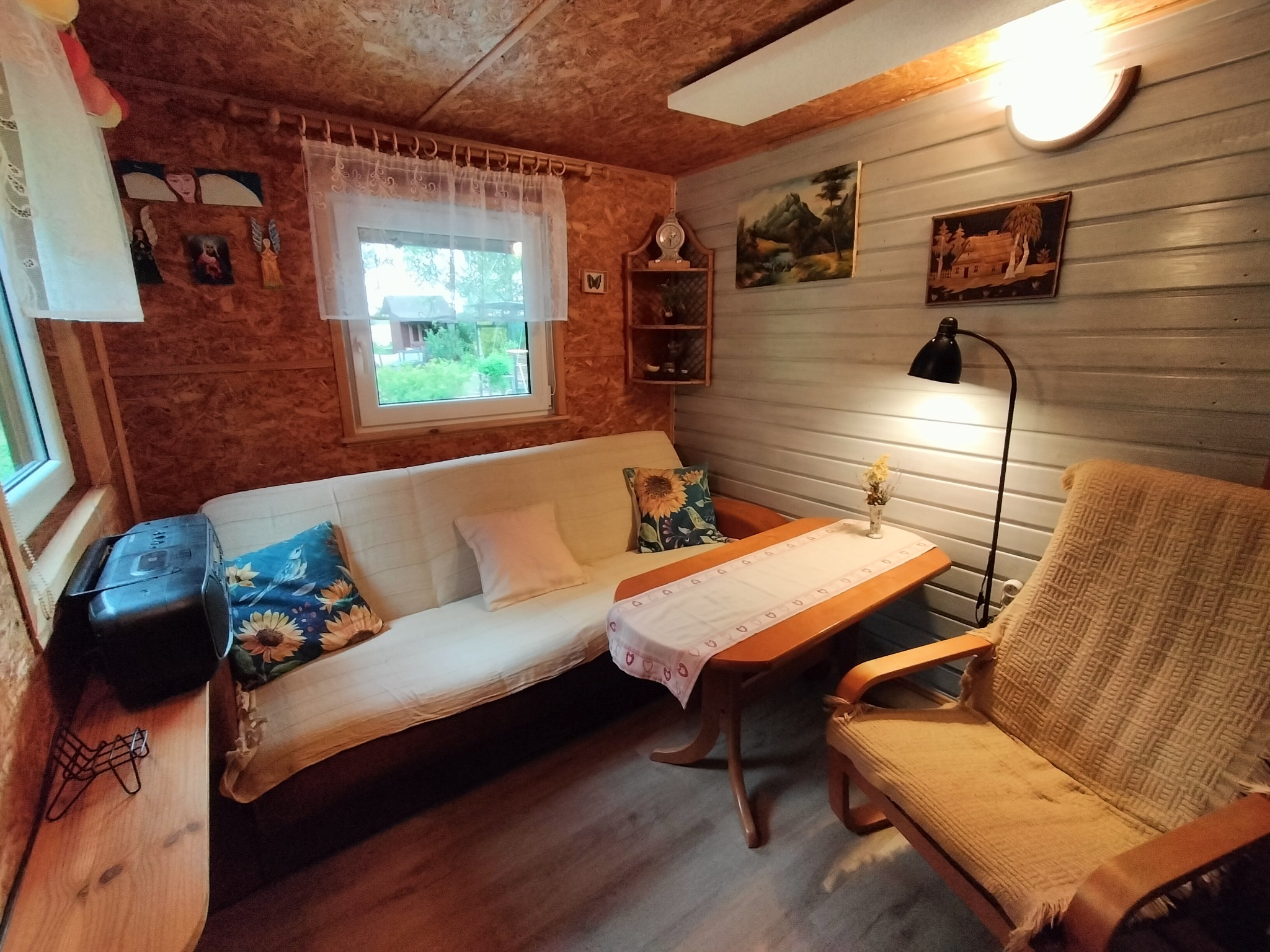 Przytulne wnętrze domku letniskowego z drewnianym wykończeniem, sofą, stolikiem i fotelem. Dekoracje ścian: obrazy i półeczka z zegarem. Okno z widokiem na zieleń.