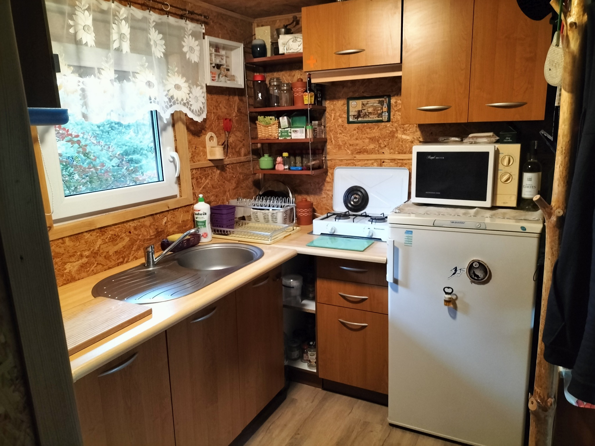 Wnętrze kuchni w domku letniskowym z drewnianymi ścianami, wyposażonego w zlew, szafki, kuchenkę, mikrofalówkę i lodówkę, okno z firanką w kwiaty.