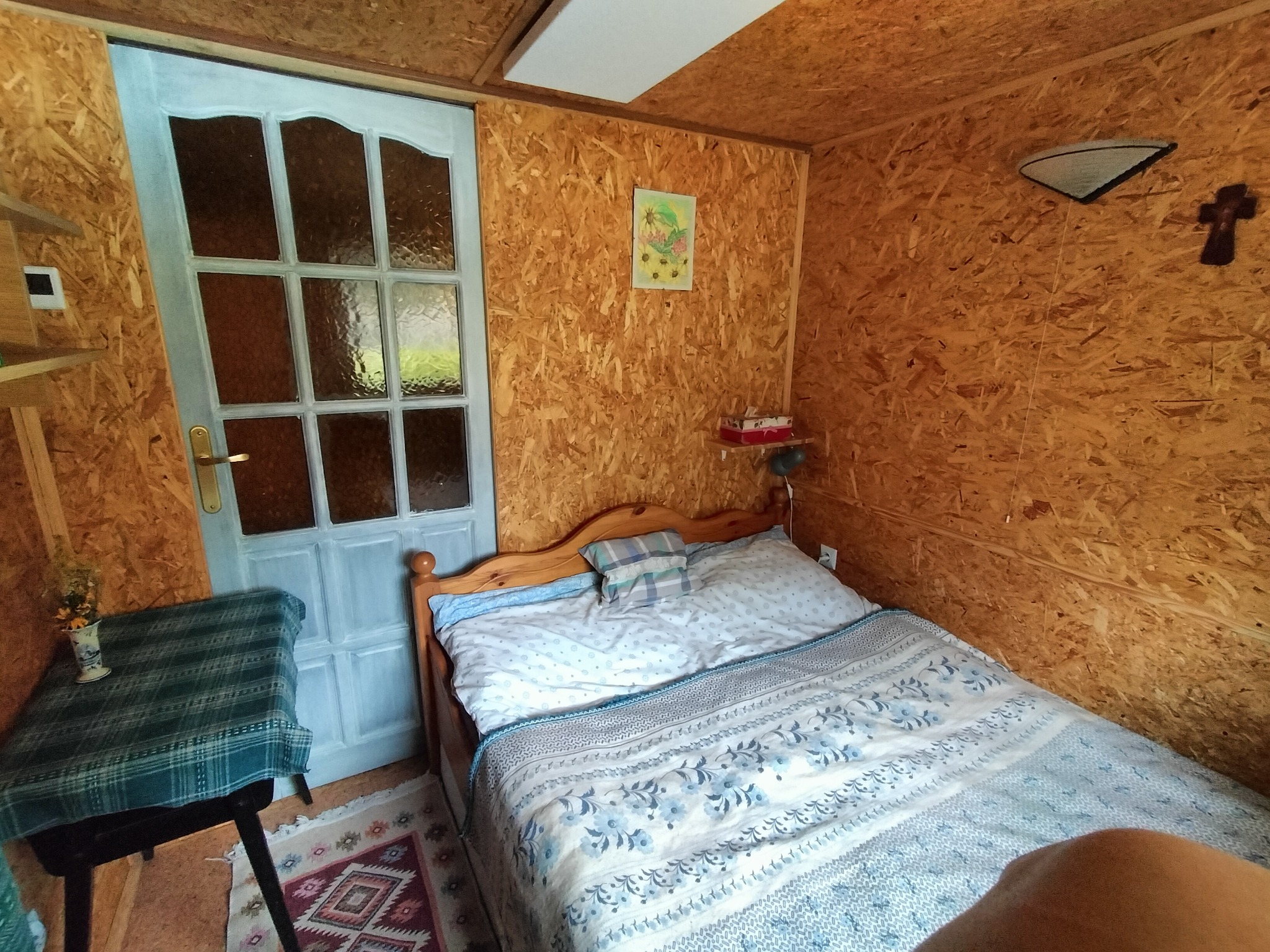 Drewniany pokój gościnny z łóżkiem dwuosobowym, stolikiem i jasnoniebieskimi drzwiami. Ściany wyłożone płytami OSB, dekoracje na ścianie. Przytulne wnętrze.