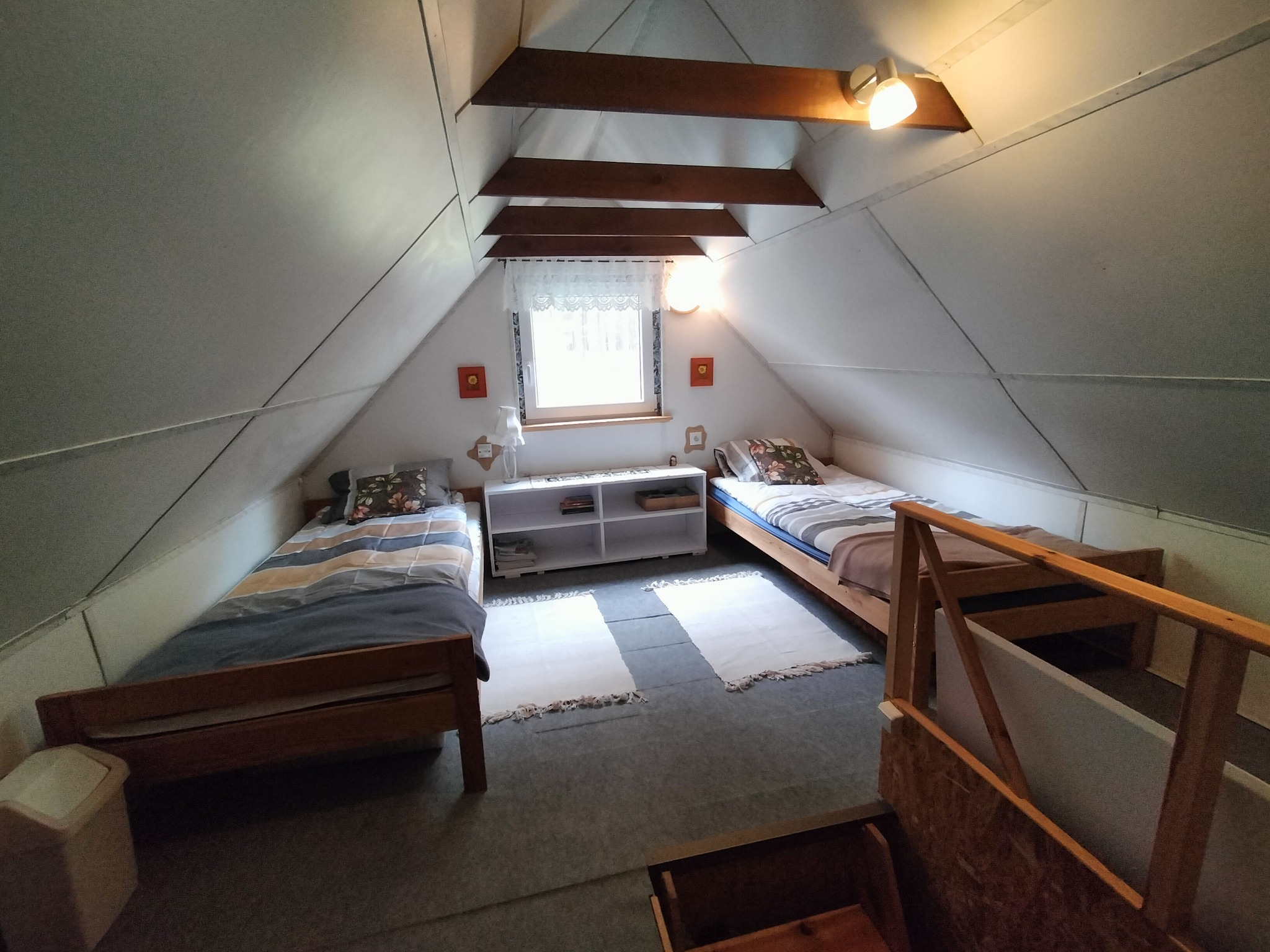 Pokój na poddaszu z dwoma pojedynczymi łóżkami, białą komodą pod oknem i drewnianymi belkami stropowymi. Ustawny pokój w pensjonacie.