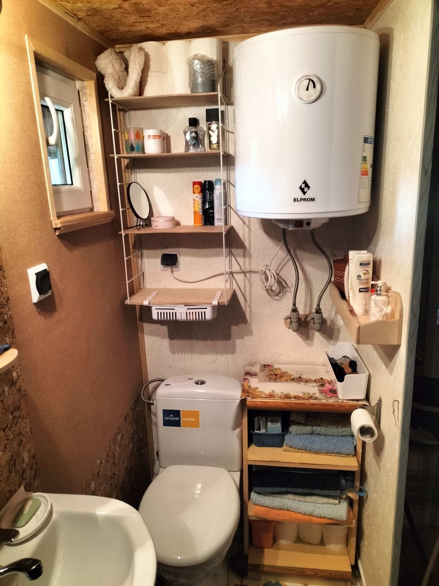 Niewielka łazienka w domku letniskowym z toaletą, umywalką, bojlerem Elprom i półkami na kosmetyki oraz ręczniki. Widoczne okno i gniazdko elektryczne.