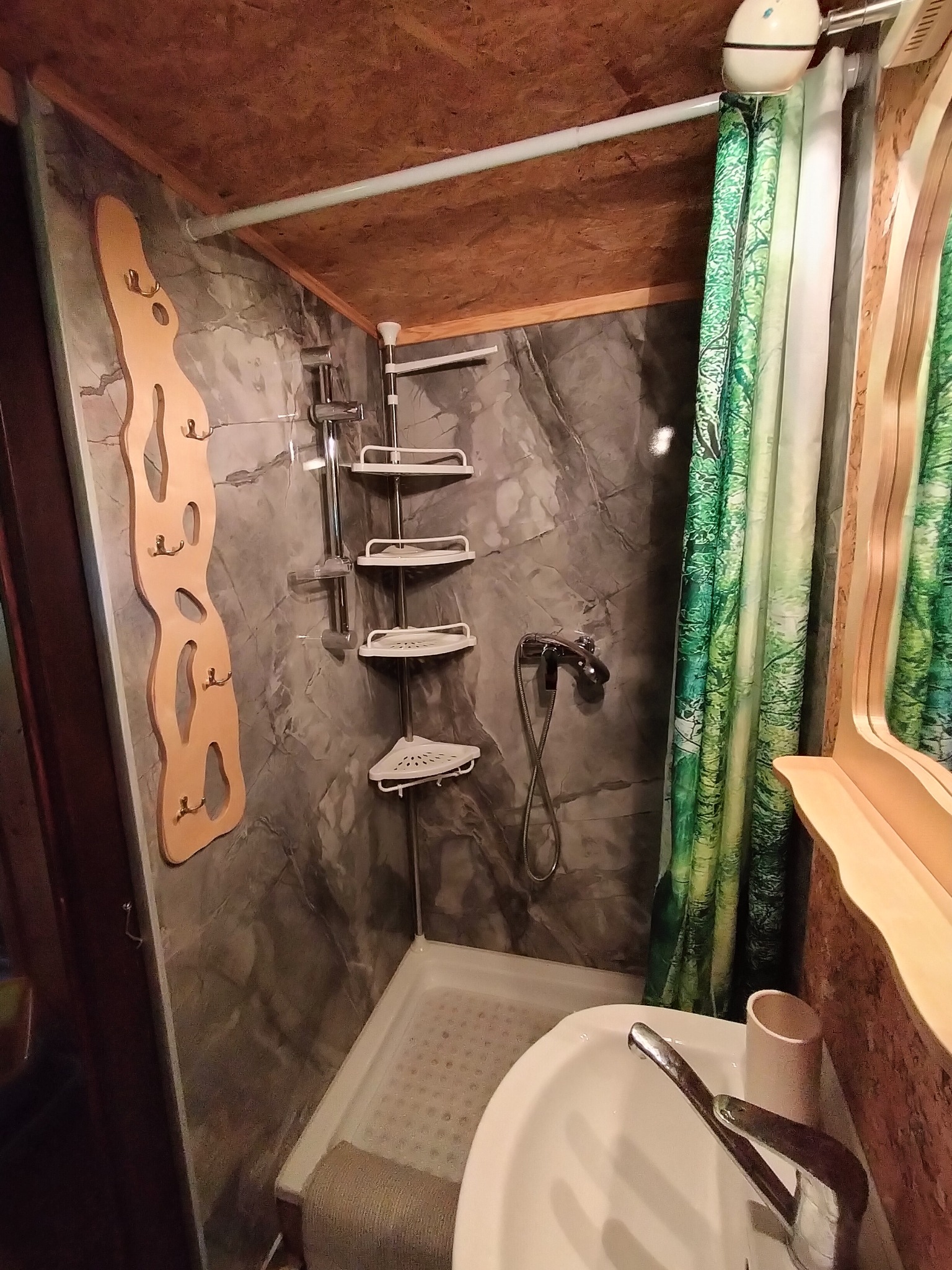 Kompaktowa łazienka w domku letniskowym: prysznic, umywalka, drewniane akcenty i zasłona z motywem leśnym. Funkcjonalne rozwiązanie przestrzeni.