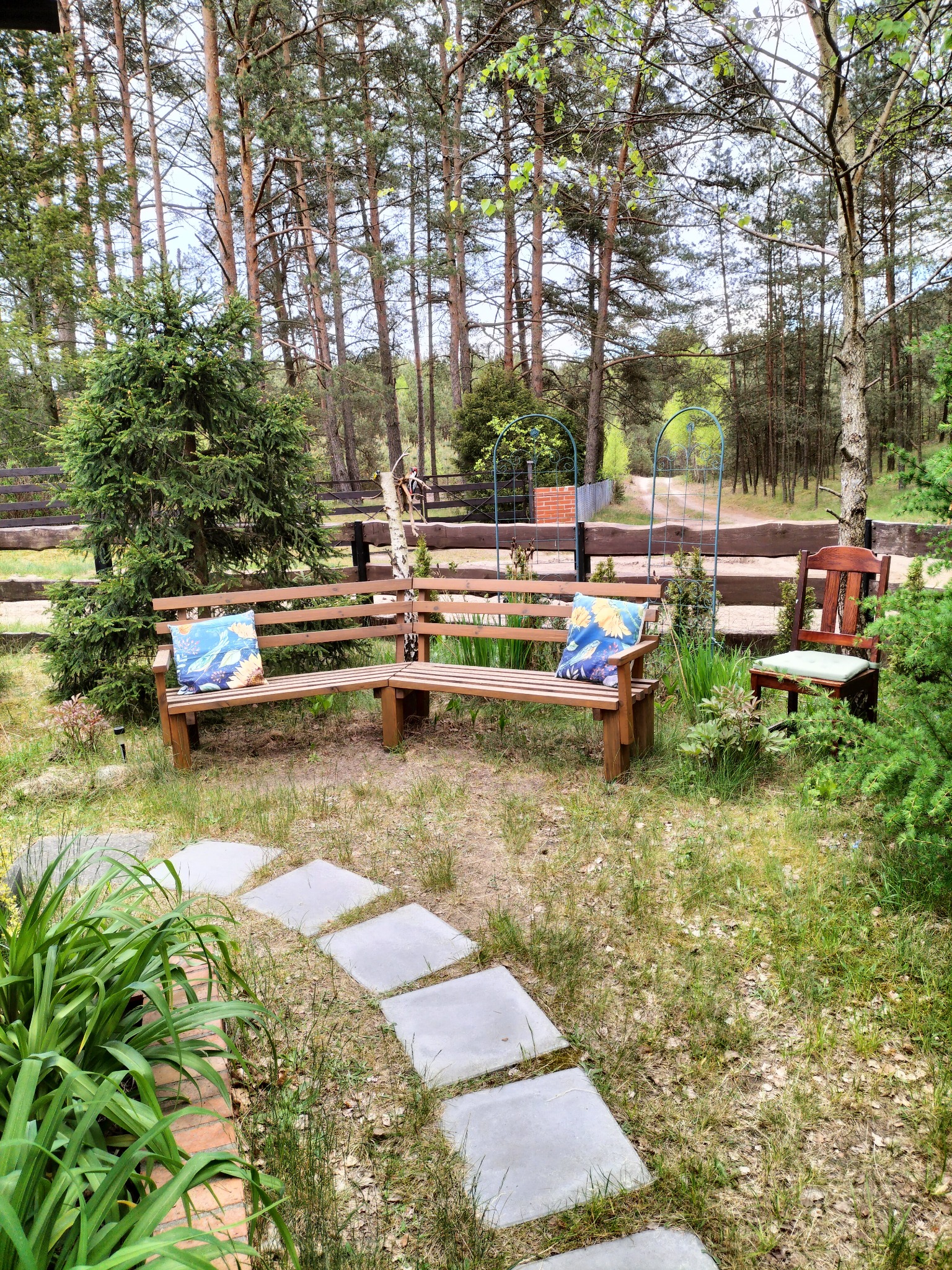 Rustykalny ogród z drewnianą ławką narożną z poduszkami i ścieżką z płyt, otoczony zielenią i drzewami. Idealne miejsce na relaks.
