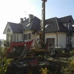 Wiercenie studni głębinowej na posesji z domem jednorodzinnym. Sprzęt wiertniczy na tle budynku z brązową dach&oacute;wką. Widoczne rury i elementy instalacji.