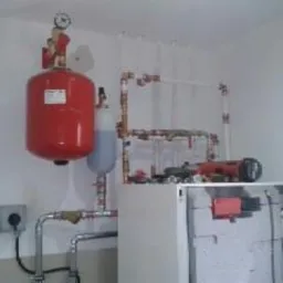 Czerwony zbiornik ciśnieniowy i instalacja grzewcza z rurami miedzianymi i plastikowymi, zamontowana w rogu pomieszczenia z białymi ścianami, widok z dołu.