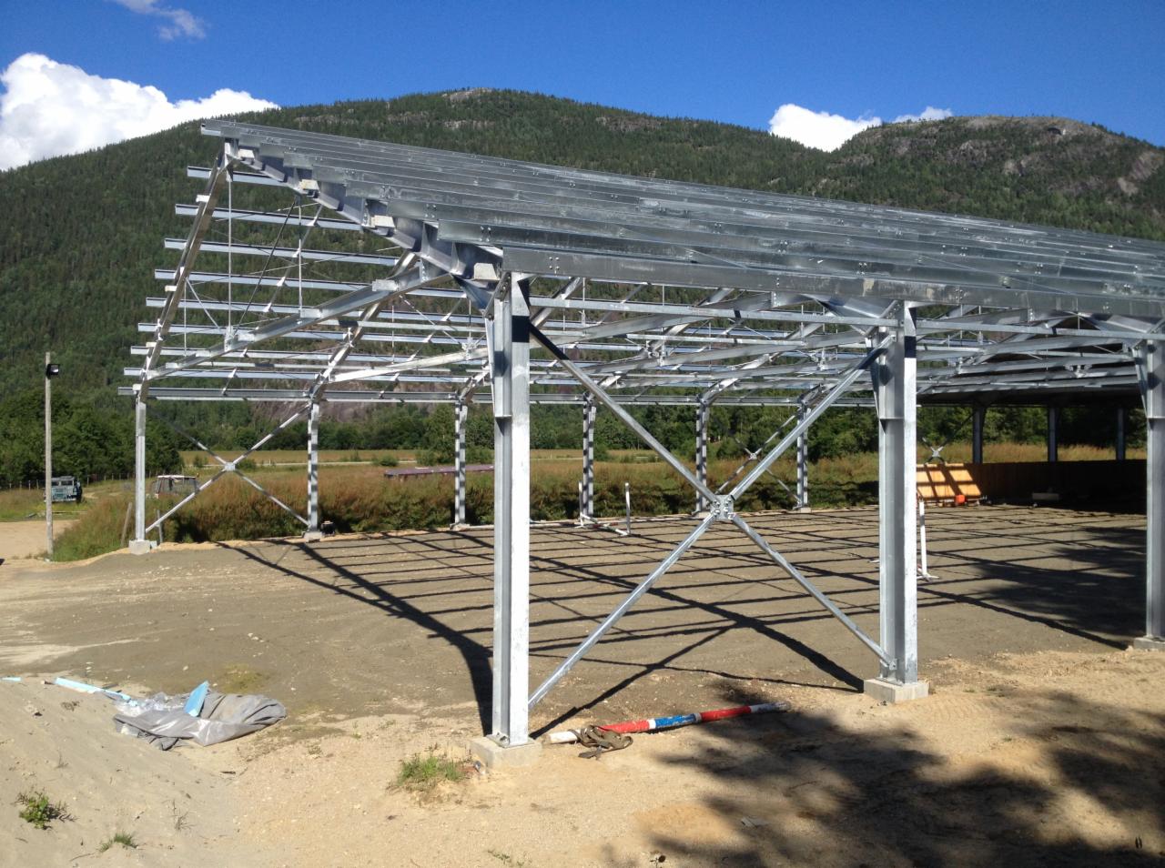 Szkielet powstającej hali z metalowych elementów na tle zielonych wzgórz i błękitnego nieba z chmurami.
