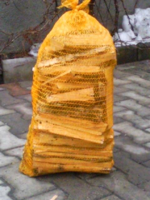 Worek z żółtej siatki wypełniony rąbanym drewnem rozpałkowym, ułożony na szarej kostce brukowej, z fragmentem ośnieżonego ogrodu w tle.