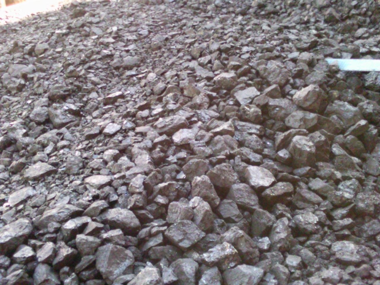 Góra ciemnego, błyszczącego węgla kamiennego o nieregularnych kształtach, z fragmentem niebieskiej rury w tle.