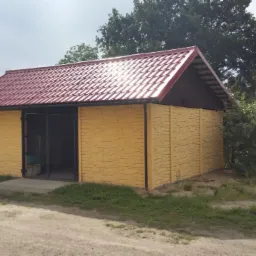 Żółty garaż z prefabrykatów betonowych, kryty czerwoną dachówką, otoczony zielenią. Widoczna brama wjazdowa i faktura paneli imitująca kamień.