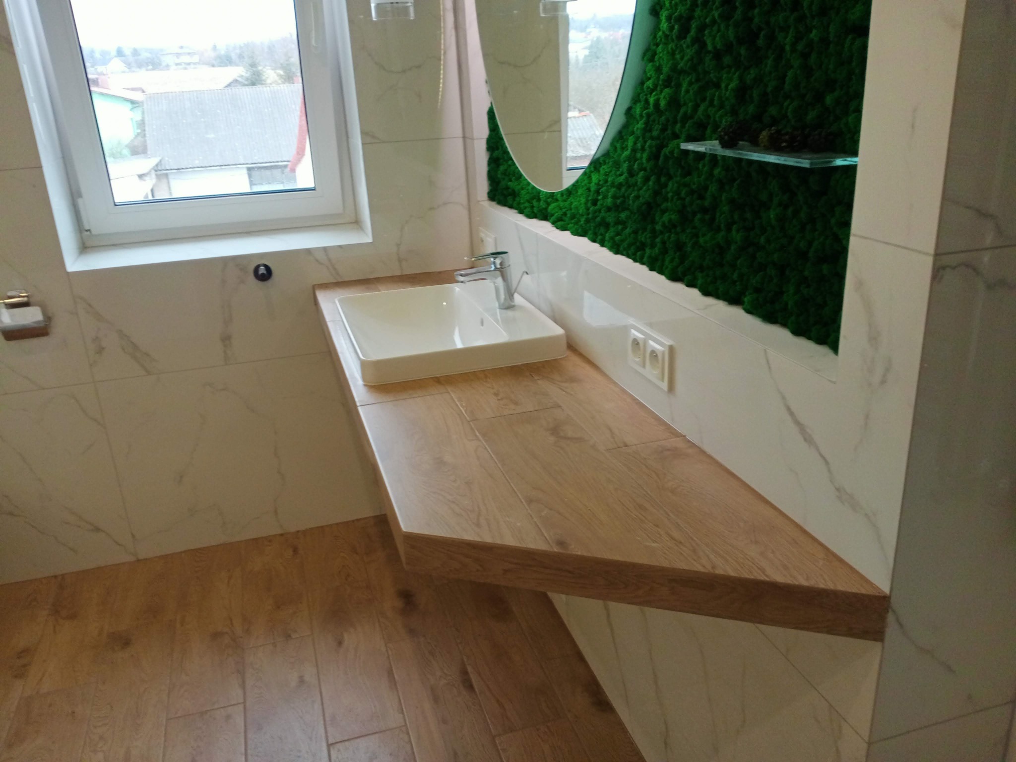 Nowoczesna łazienka z drewnianym blatem pod umywalką, białymi płytkami imitującymi marmur na ścianach i podłodze oraz ścianą z mchu dekoracyjnego za lustrem.