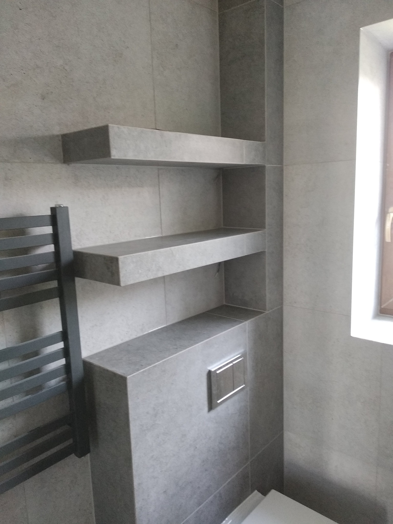 Wnętrze łazienki wykończone szarą płytką ceramiczną z wbudowanymi półkami i grzejnikiem drabinkowym, widoczne elementy podtynkowej instalacji WC.