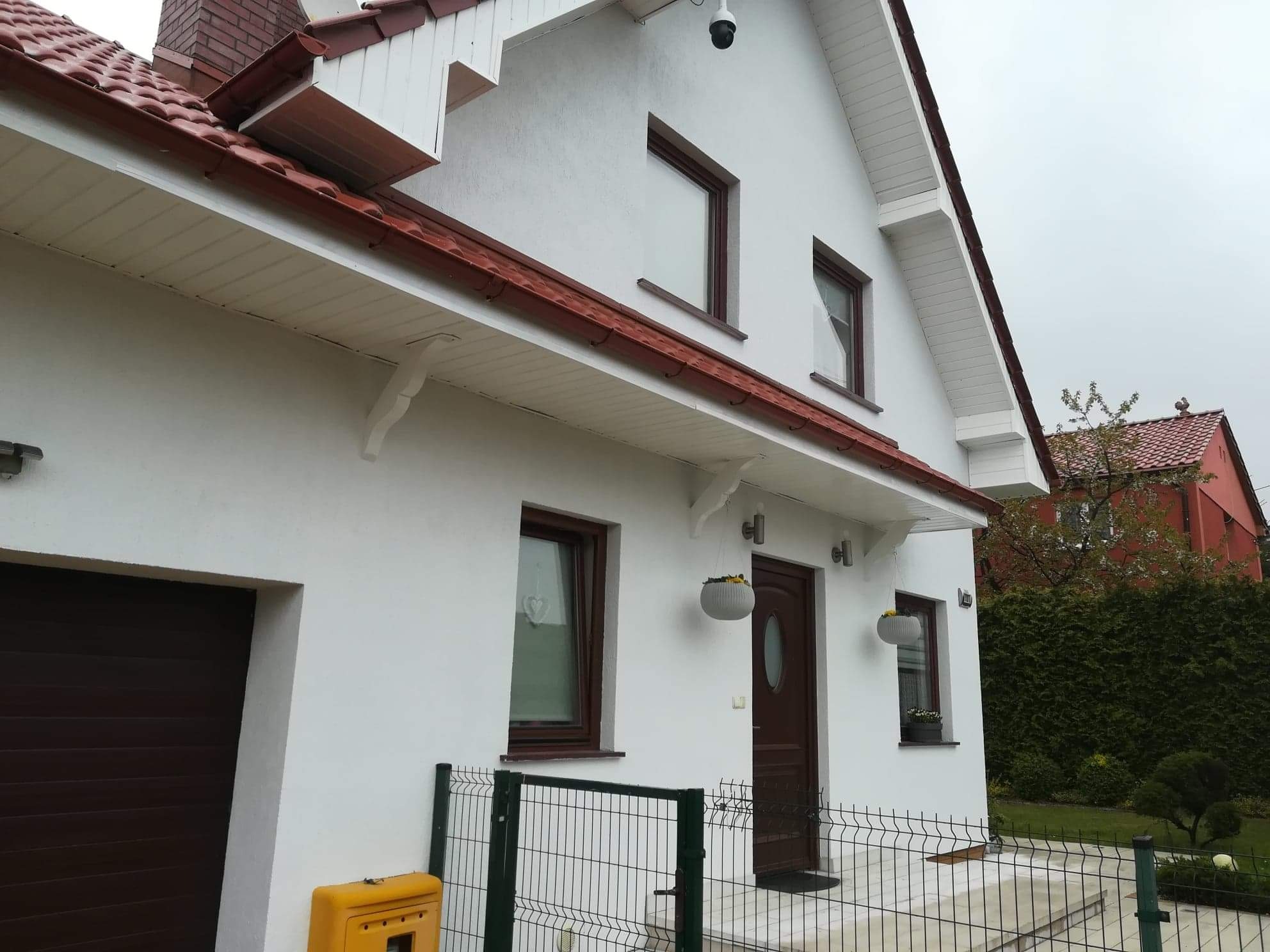 Biały dom z brązowymi oknami i dachem z czerwonej dachówki, widoczny zza zielonego ogrodzenia, z elementami dekoracyjnymi takimi jak wiszące doniczki i lampy zewnętrzne.