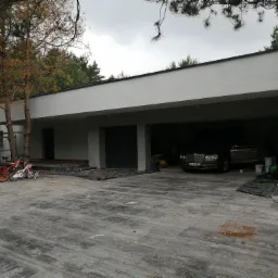 Nowoczesny dom jednorodzinny z garażem w trakcie prac wykończeniowych elewacji; widoczny pracownik na rusztowaniu, zabezpieczona folią podłoga i dwa luksusowe samochody w garażu.