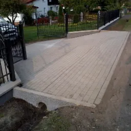 Ułożona kostka brukowa na podjeździe z czarnym ogrodzeniem i bramą. Widoczny fragment ulicy i samochodu. Zakończony drenaż wody.