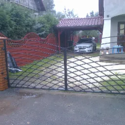 Czarna, metalowa brama wjazdowa z ceglanymi słupkami na posesji. W tle czerwone ogrodzenie, wiata garażowa i biały samochód. Asfaltowa droga w deszczu.