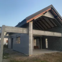 Surowy stan domu jednorodzinnego z bloczków z keramzytu, widoczna konstrukcja dachu z czarną blachodachówką i betonowe słupy nośne, budowa w trakcie, poranne światło.