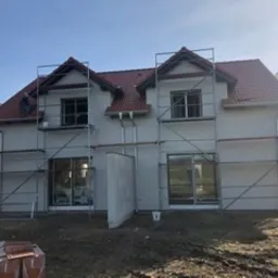 Dwukondygnacyjny dom w trakcie prac elewacyjnych, z widocznymi rusztowaniami, świeżo otynkowaną fasadą i pracownikiem na dachu. Czerwona dachówka, szare ściany, błękitne niebo w tle.