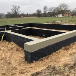 Zalane betonem fundamenty domu jednorodzinnego z hydroizolacją w postaci czarnej folii kubełkowej, widok na teren budowy z domami w tle.