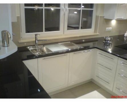 Fragment kremowej zabudowy kuchennej z czarnym blatem, widoczny zlew z baterią pod oknem i narożna szafka z szufladami.