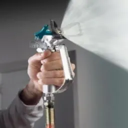 Pracownik w masce ochronnej aplikuje farbę natryskowo na ścianę, widoczny pistolet natryskowy i rozpylona farba.