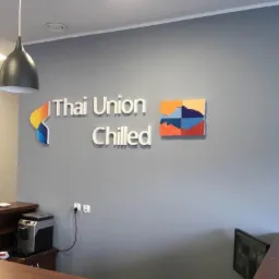 Wnętrze biura z logo firmy Thai Union Chilled na szarej ścianie, oświetlone lampami wiszącymi, obok ekspres do kawy.