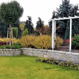 Ujęcie ogrodu z trawnikiem, kamiennym murkiem, rabatami z różnorodnymi roślinami, drzewami, pergolą i działającym zraszaczem.