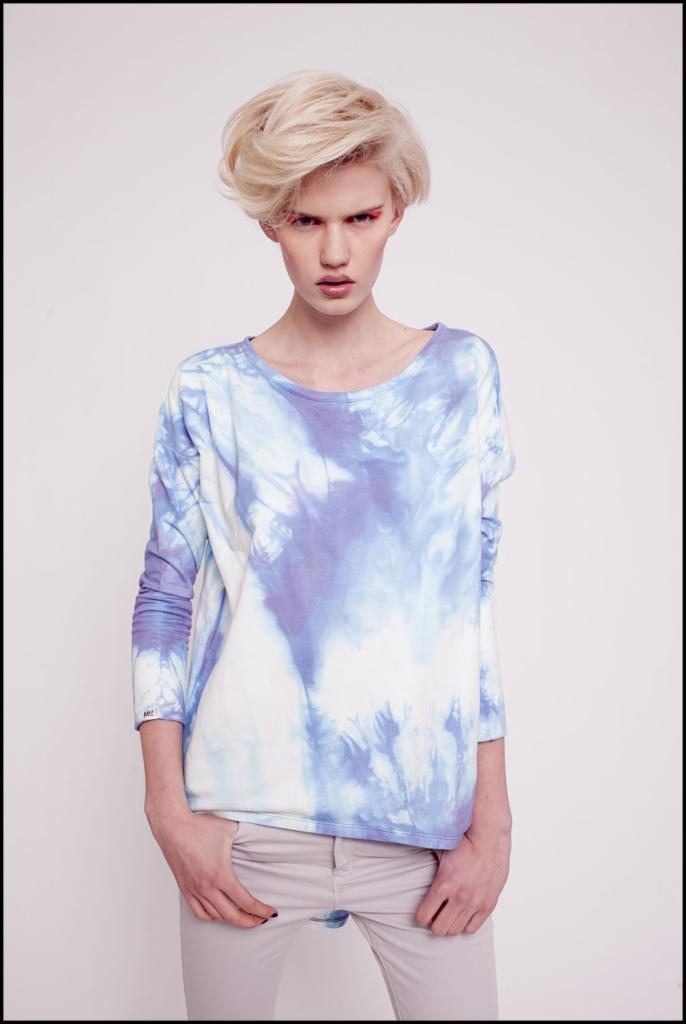 Modelka z krótkimi blond włosami prezentuje bawełnianą bluzę ICE MIRROR z efektem tie-dye w odcieniach błękitu i fioletu na białym tle, z rękami włożonymi w kieszenie szarych spodni.