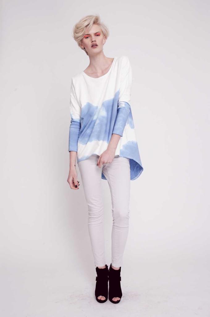 Modelka prezentuje bawełnianą bluzę Blue Cloud z efektem tie-dye, białe spodnie i czarne botki na białym tle.