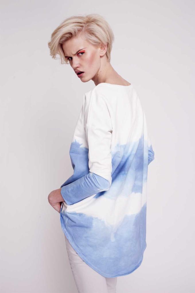 Kobieta z krótkimi blond włosami, odwrócona przez ramię, prezentuje białą bluzę z długim rękawem i efektem ombre w odcieniach błękitu, na białym tle.
