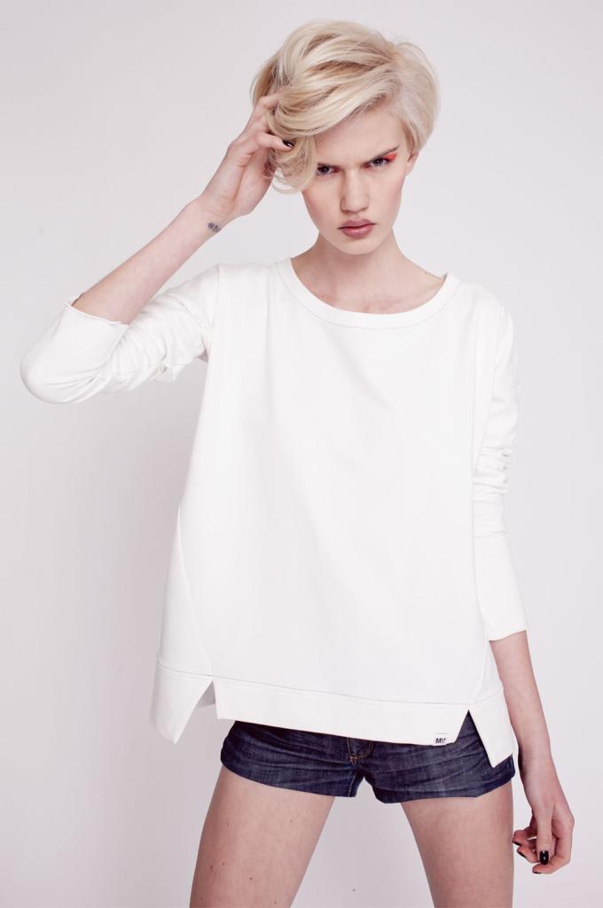 Modelka z krótkimi blond włosami, w białej bawełnianej bluzie z długim rękawem i asymetrycznym dołem, pozuje na białym tle, dotykając włosów i prezentując tatuaż na nadgarstku.