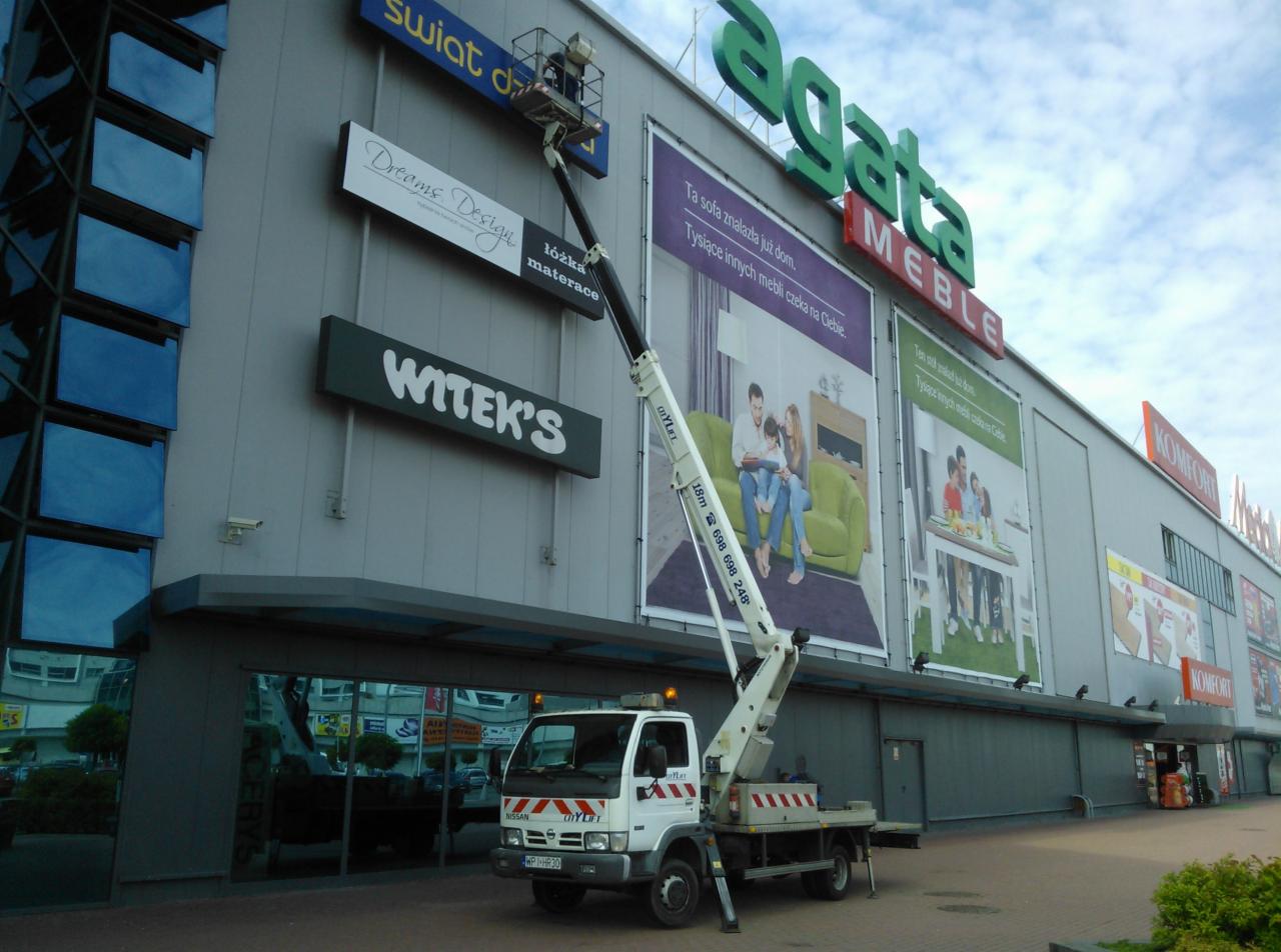 Montaż reklam zewnętrznych na fasadzie budynku Agata Meble z użyciem podnośnika koszowego CityLift, widoczny pracownik w koszu i reklamy Witek's, Dreams Design, Komfor oraz inne elementy...