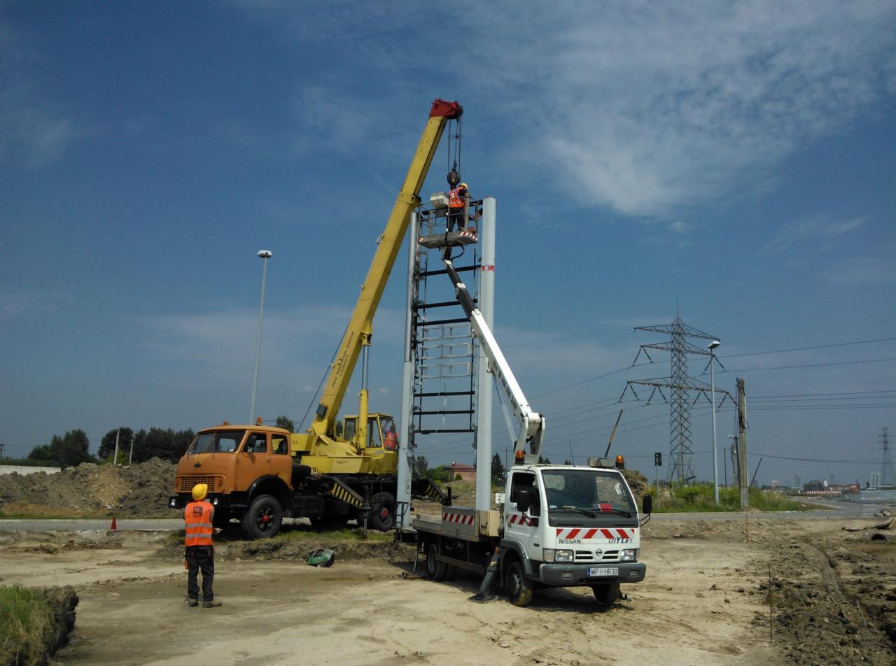 Dwóch pracowników w pomarańczowych kamizelkach na platformie podnoszonej przez żółty dźwig, obok stoi biały samochód dostawczy z wysięgnikiem, w tle słupy wysokiego napięcia i błękitne niebo.