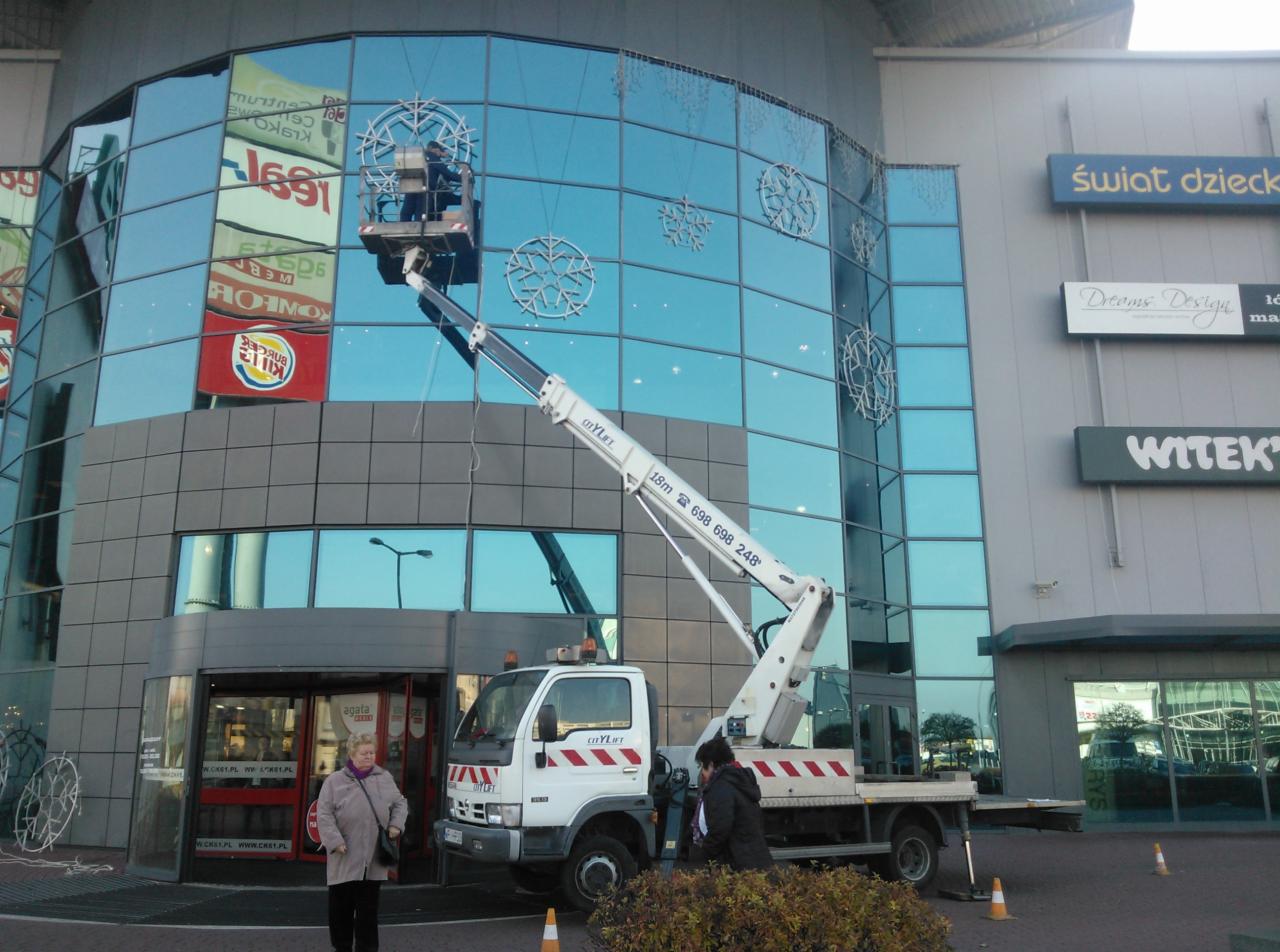 Montaż świątecznych dekoracji w postaci śnieżynek na szklanej fasadzie centrum handlowego przy użyciu białego podnośnika koszowego CityLift, widoczni pracownicy w koszu i na ziemi,...