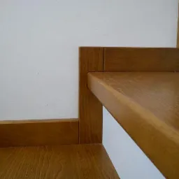 Fragment schod&oacute;w obłożonych drewnem o ciepłym odcieniu, widoczne połączenie stopnia z pionową częścią schod&oacute;w, z wyraźnym usłojeniem drewna i gładkim wykończeniem, na tle białej ściany.