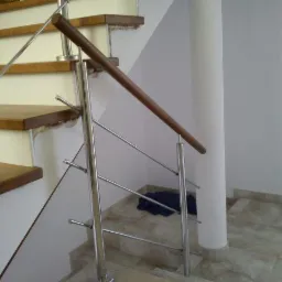 Stalowa balustrada z drewnianym pochwytem zamontowana przy schodach z drewnianymi stopniami, widoczny fragment kolumny i podłogi wyłożonej płytkami.