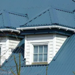 Niebieski dach z blachy falistej z białymi oknami lukarn i systemem rynnowym, widok z dołu, gałęzie drzew na pierwszym planie.