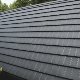 Szary dach z blachy o trapezoidalnym profilu, widok z dołu, z fragmentem zieleni w tle, ukazujący regularny wzór i fakturę pokrycia.