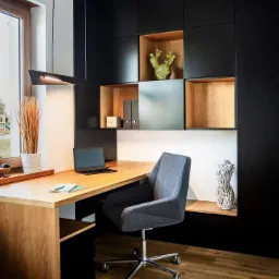 Nowoczesne biuro z drewnianym biurkiem, szarą tapicerowaną krzesłem, czarnymi szafkami w zabudowie i dekoracyjnymi elementami na półkach.