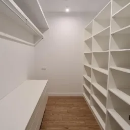 Wnętrze garderoby z białymi półkami i drewnianą podłogą ułożoną w jodełkę, oświetlone punktowymi lampami sufitowymi.