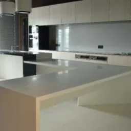 Biały, nowoczesny blat kuchenny w kształcie litery U, z widocznym fragmentem czarnego blatu wyspy kuchennej i jasnymi szafkami w tle, oświetlony przez wiszące lampy.