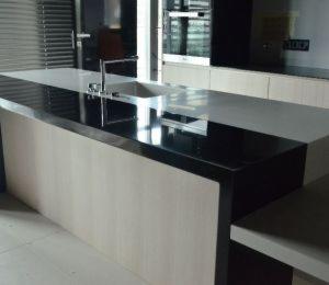 Wyspa kuchenna z blatem w kolorze czarnym i białym, z nowoczesną baterią, z frontami w odcieniach beżu i szarości, widoczna w jasnym wnętrzu kuchni.