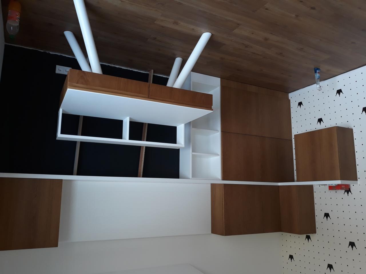 Częściowo zamontowana szafa wnękowa z białymi półkami i drewnianymi frontami na tle czarnej ściany i tapety w korony.