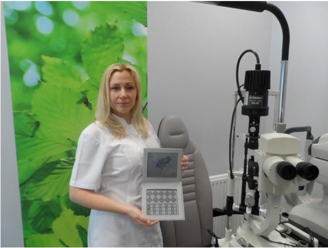 Lekarz w białym uniformie prezentuje tablicę do badania wzroku z bliska, w tle widoczny biomikroskop okulistyczny i dekoracyjna grafika liści.