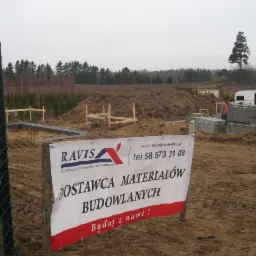 Fundamenty domu w trakcie budowy, widoczny baner reklamowy dostawcy materiałów budowlanych, w tle sterta ziemi, bloczki betonowe i przyczepa kempingowa.